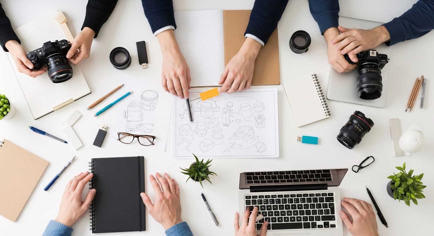 A working desk with cameras, printed sketches and open laptops laid out mid-project.